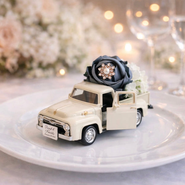 Decorative model of a vintage truck with flowers on a white plate, set against a blurred background of candles and glasses.