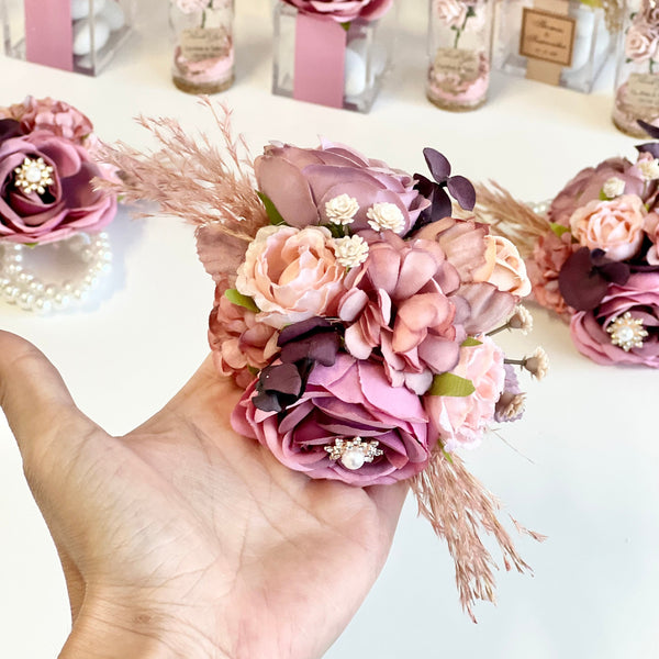 Hand holding a bouquet of pink and purple flowers with decorative elements on a white background.
