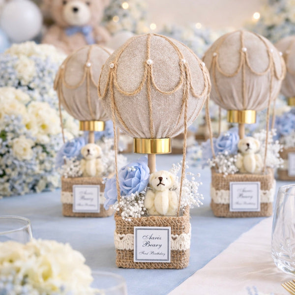 Decorative hot air balloon centerpieces on a table with flowers and teddy bears, set against a blurred background with 'Baby' sign.