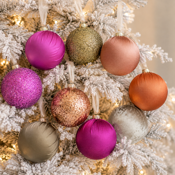 Nine colorful Christmas tree ornaments in various textures (matte, glitter, velvet) in pink, gold, bronze, and silver, hanging on a white-flocked Christmas tree with warm string lights.