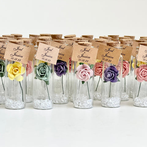 Small glass bottles with colorful paper flowers and name tags on a white background