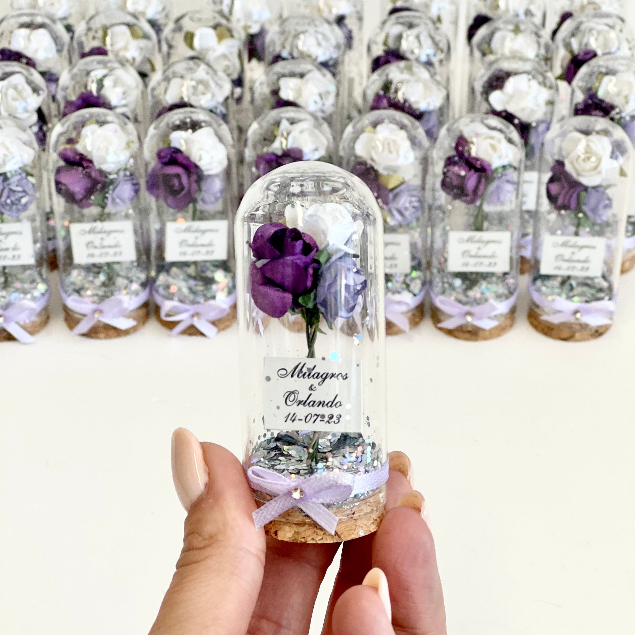 Hand holding a small glass bottle with a purple flower inside, surrounded by similar bottles on a white background.