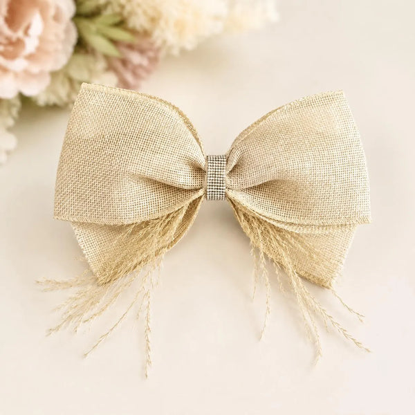 A large decorative hair bow made from textured beige burlap fabric with frayed fringe on the sides, centered on a soft cream background with blurred pale flowers in the upper left.