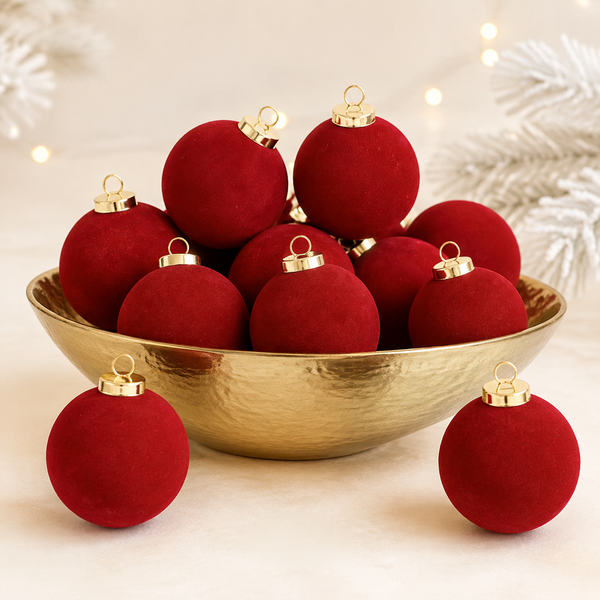 A set of deep red velvet Christmas ornaments with gold caps, displayed in a hammered gold bowl with two ornaments resting in front on a light beige surface, decorated with soft white pine branches and warm bokeh lights in the background.
