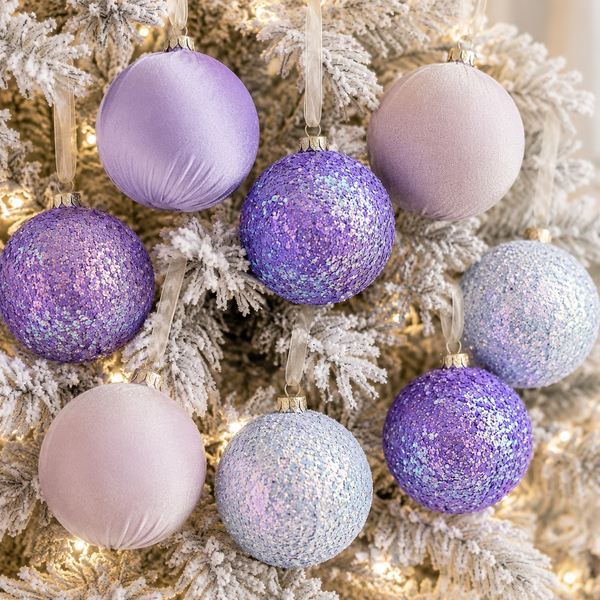 A set of eight Christmas tree ornaments in pastel lavender, lilac, and light blue, including both glittery and satin finishes, hanging on a white-flocked Christmas tree with warm string lights.
