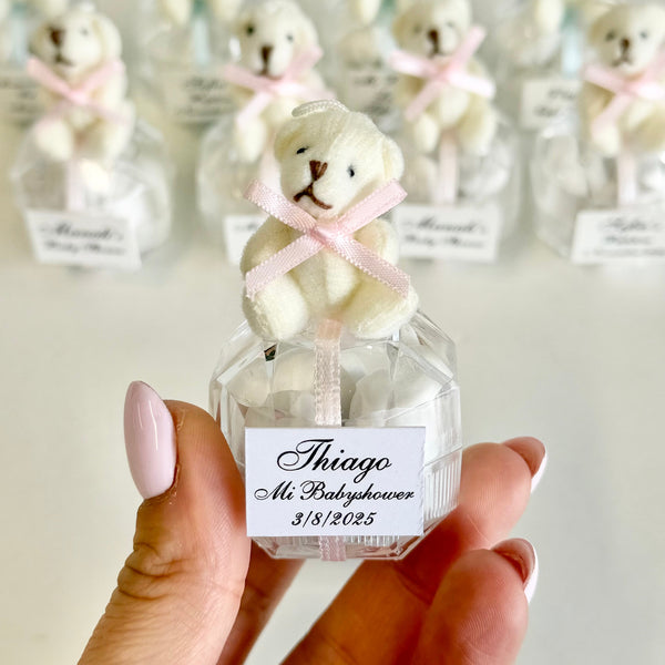 Small bottle with a teddy bear toy and personalized label held by a hand, with more bottles in the background.