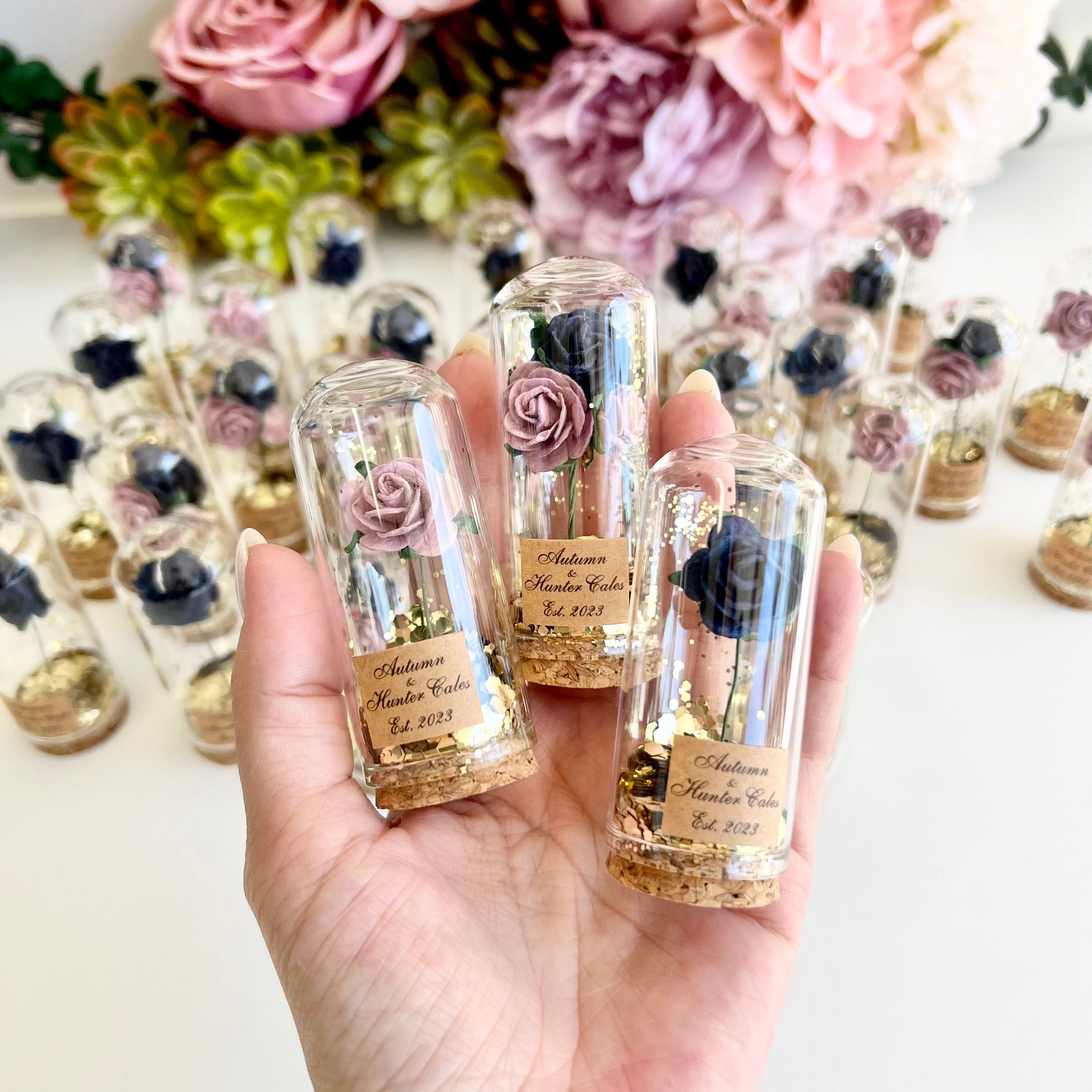 Three small transparent bottles with floral decorations held by a hand, with more bottles and flowers in the background.
