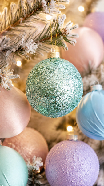 A close-up of a glittery mint green Christmas ornament hanging on a frosted pine tree branch, surrounded by pastel pink, blue, and lavender ornaments with warm string lights in the background.