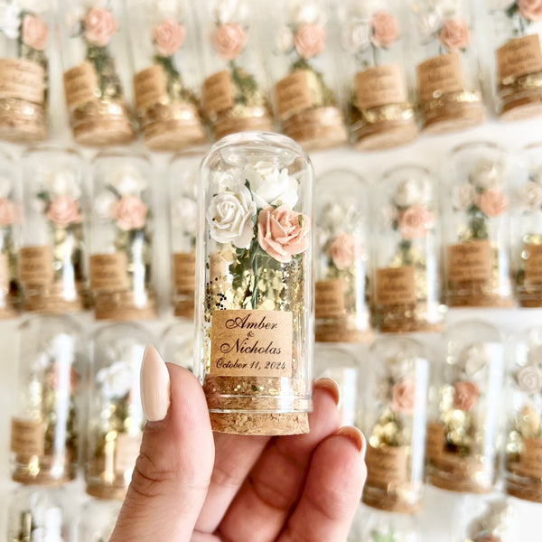 Hand holding a small glass bottle with floral arrangement against a wall of similar bottles.