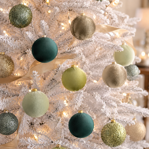 A close-up of a white frosted Christmas tree decorated with matte and glitter ornaments in sage green, teal, champagne, and gold, with warm string lights in the background.