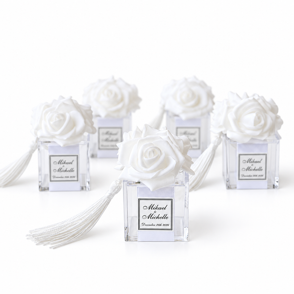 Five white rose favor bags in clear square glass containers with white tassels, each labeled with personalized names on a clean white background.