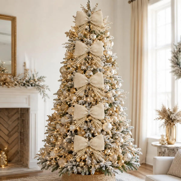 A decorated Christmas tree in a bright, neutral-toned living room. The tree is frosted with white snow-like dusting and adorned with large beige burlap bows, white and gold ornaments, and warm string lights.