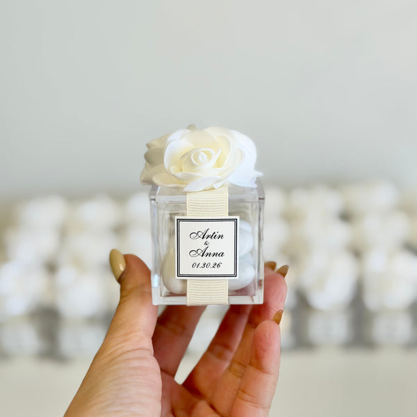 Hand holding a small perfume bottle with a white rose on top against a blurred background