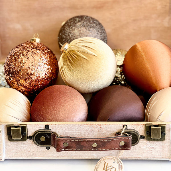 A luxurious set of handmade Christmas ornaments in warm tones of copper, bronze, chocolate brown, champagne, and gold. The ornaments, crafted from materials like glitter, satin, and velvet, are elegantly arranged inside a vintage-style wooden suitcase with a leather handle and a branded WhiteRoom tag. The festive color palette and texture variety create a rich, cozy, and sophisticated holiday decor aesthetic.