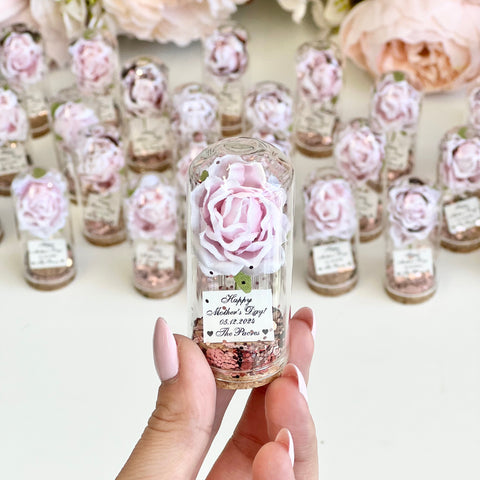 Small glass bottle with a preserved pink rose held by a hand, with more bottles and flowers in the background.