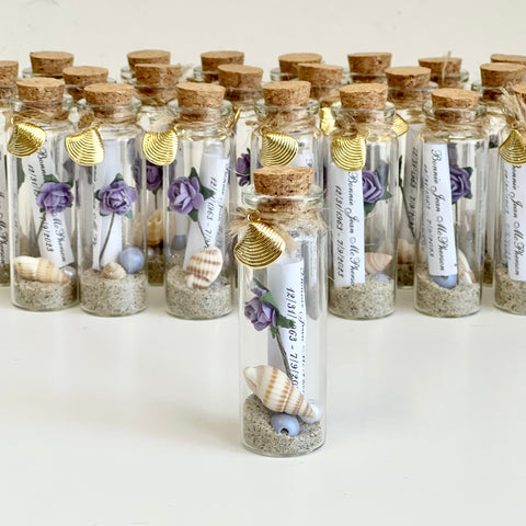 Small glass bottles with cork lids, containing decorative elements like sand, shells, and small plants, on a white background.