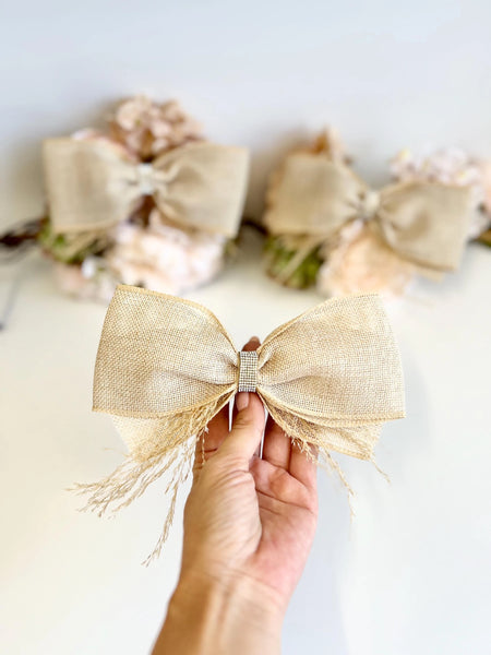 A hand holding a large beige burlap bow with frayed edges and a small rhinestone-embellished center, with blurred matching bows in the background.