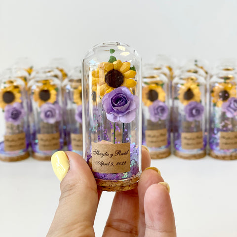 Glass bottle with preserved flowers held by a hand, with more bottles in the background.