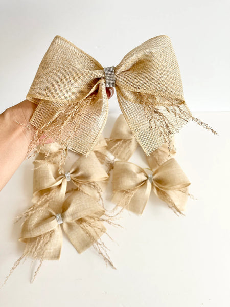 A hand holds a large beige burlap bow with dried grass accents, above several smaller matching burlap bows with rhinestone center details, on a plain white background.