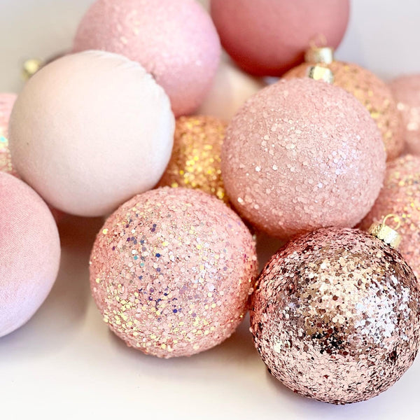 A close-up pile of round decorative ornaments in soft pink, blush, and rose-gold tones, including matte, glitter, and metallic finishes on a light neutral background.