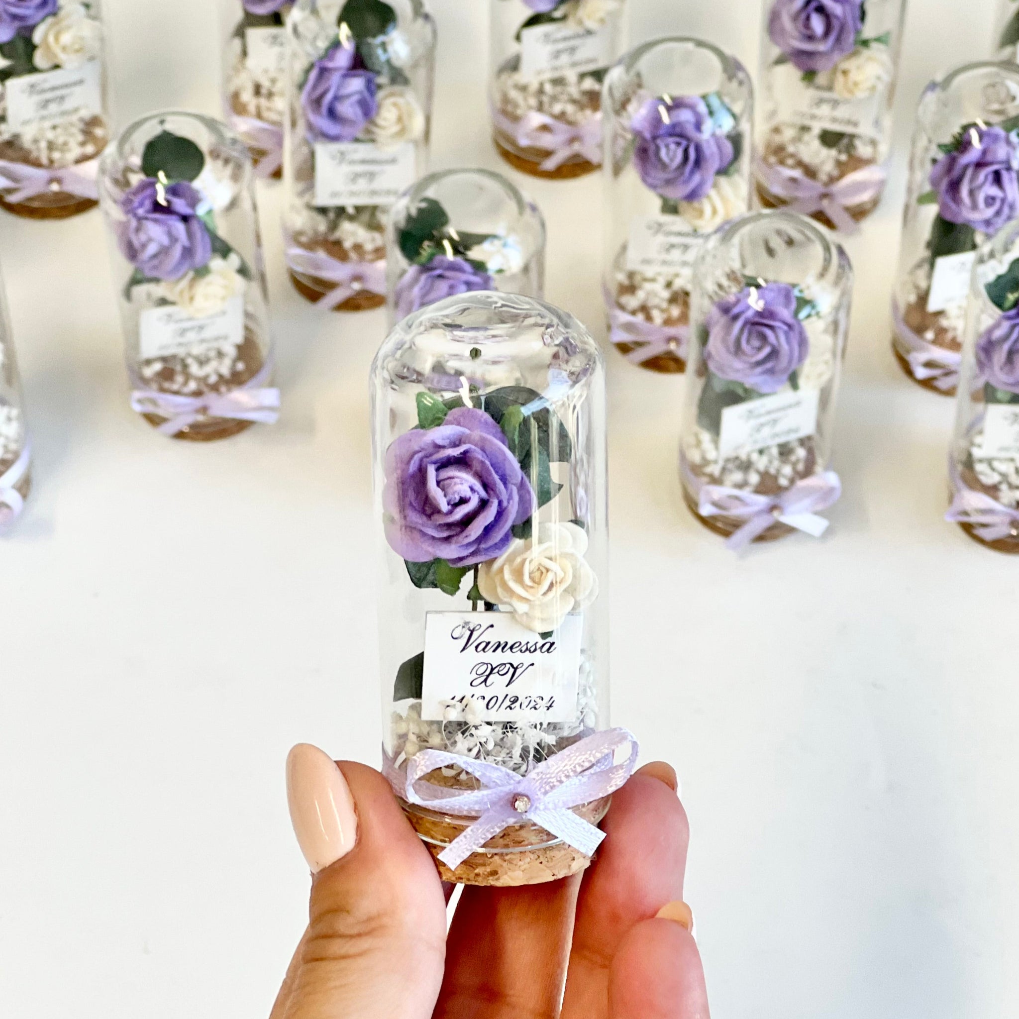 Hand holding a small glass bottle with a preserved purple flower against a white background with more similar bottles.