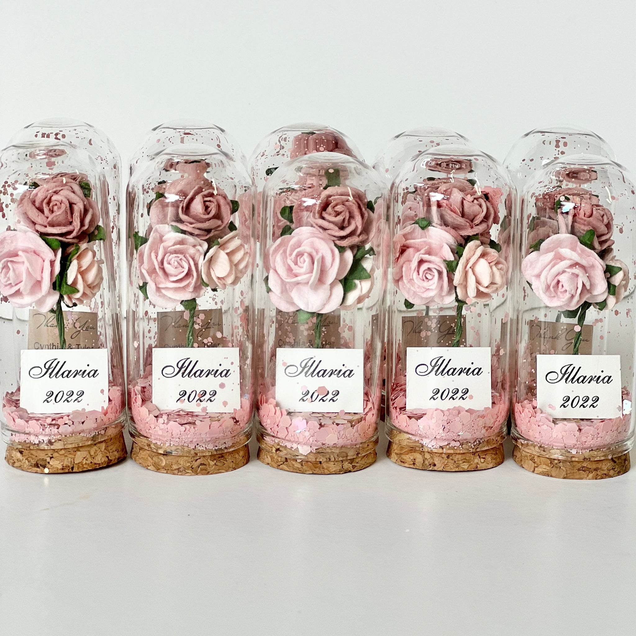 Five glass jars with preserved pink roses and 'Ilaria 2022' labels on a white background