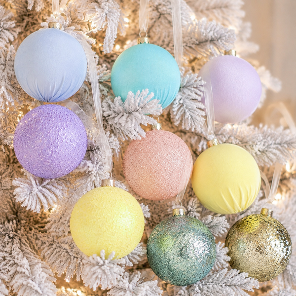 Nine pastel Christmas tree ornaments in various textures—smooth, glitter, and sparkly—are hanging on a flocked white Christmas tree with warm string lights.