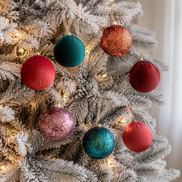 Six Christmas tree ornaments in red, teal, and glittery pink and copper colors hanging on a flocked white Christmas tree with warm string lights.