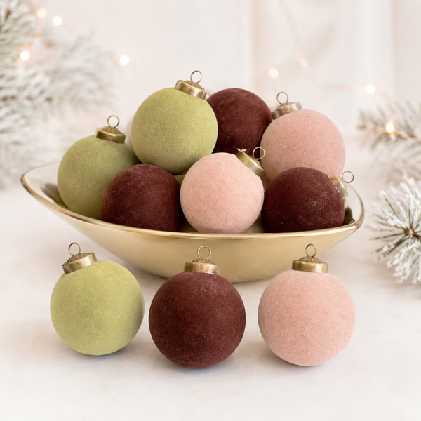 Six round Christmas ornaments in muted sage green, dusty rose, and burgundy, displayed in a gold bowl with three more placed in front on a white surface.