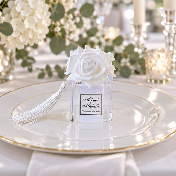 A personalized clear acrylic favor box with a white fabric rose and tassel, displayed on a white and gold charger plate at a beautifully set wedding table.