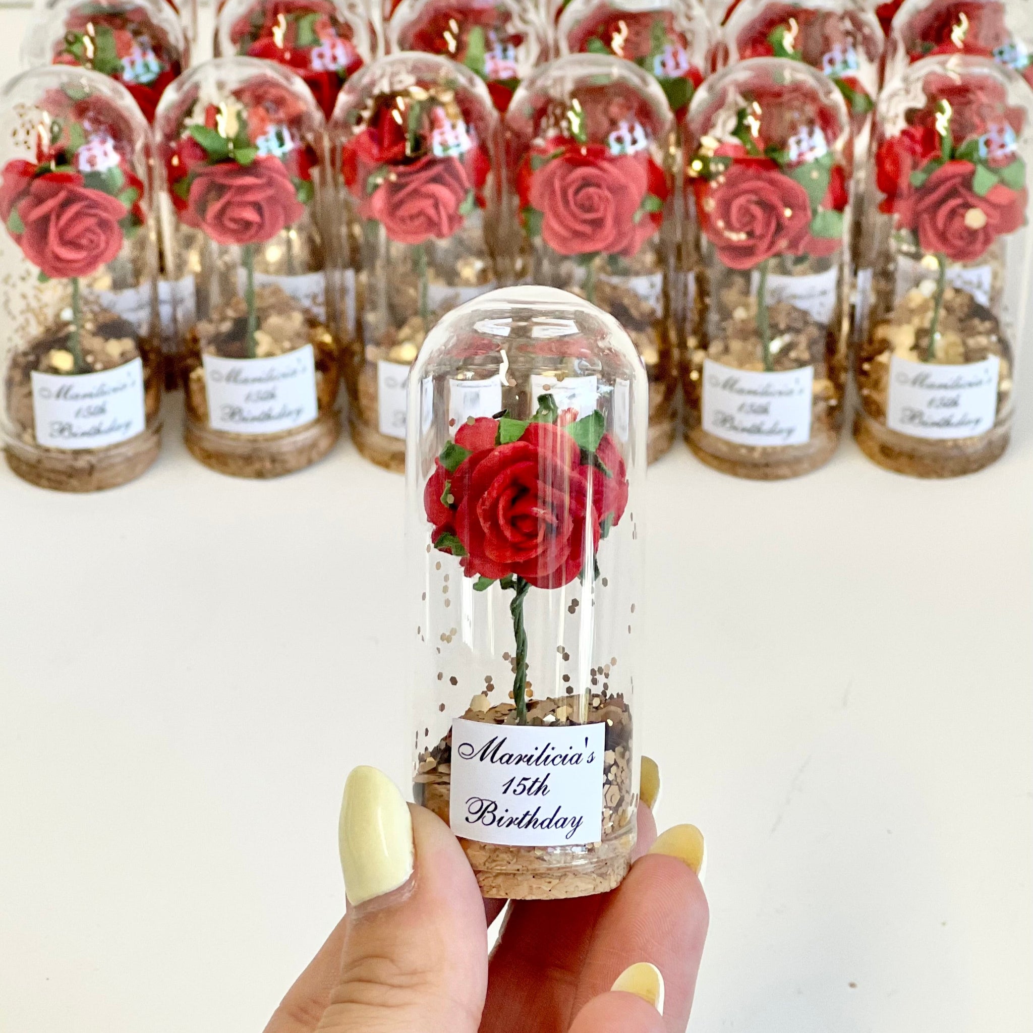 Hand holding a small glass bottle with a red rose, surrounded by more bottles with roses on a white background.