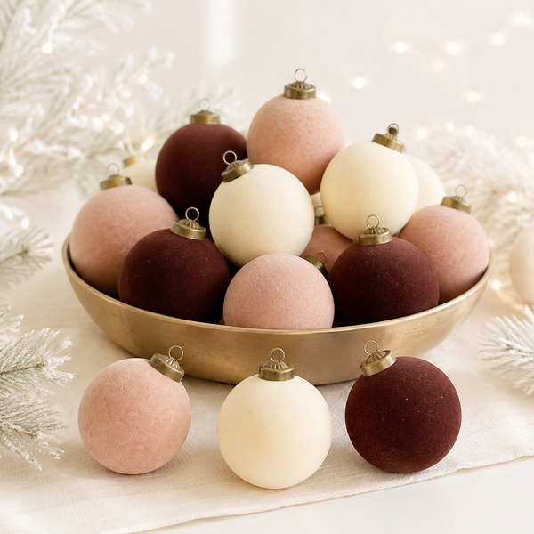 A collection of matte velvet Christmas ornaments in dusty pink, cream, and deep burgundy, arranged in a gold bowl with a few placed in front on a white fabric surface with festive white branches and soft lights in the background.