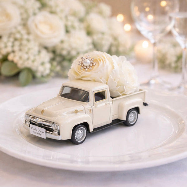 Small model truck with flowers on a white plate, set against a blurred floral and candle background.