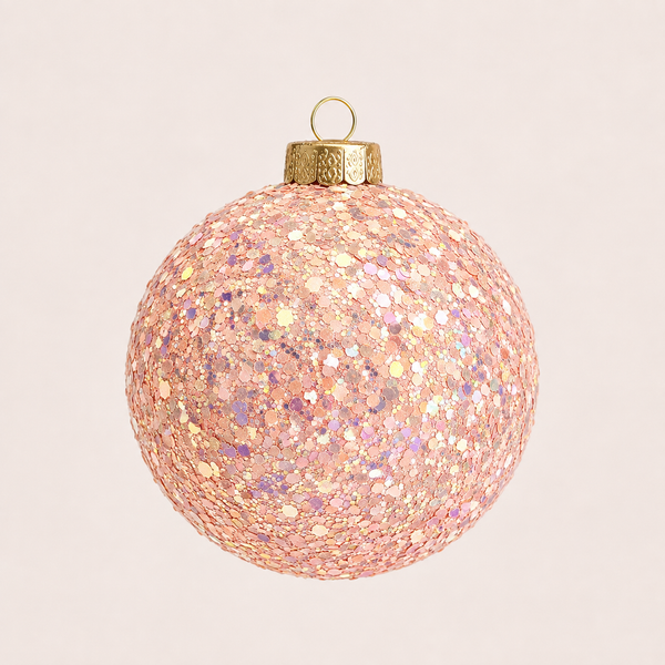 A spherical Christmas ornament covered in shimmering rose gold and pink glitter, featuring a textured gold cap and hanging loop, set against a solid pale pink background.