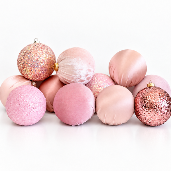 A group of decorative round ornaments in soft pink and rose gold tones, including glittery, velvet, and satin finishes, arranged on a white surface against a white background.