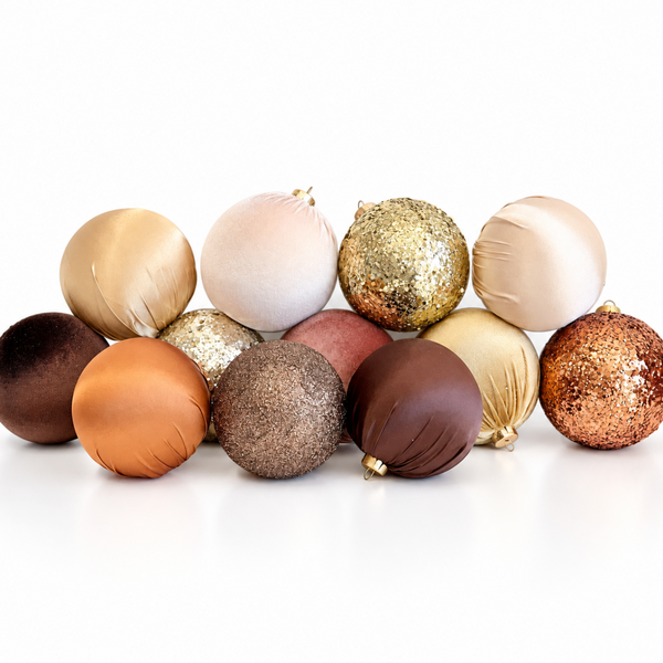 A group of twelve assorted Christmas ornaments in various colors and textures, including matte, satin, velvet, glitter, and ribbed finishes in shades of gold, bronze, copper, brown, pink, and cream, arranged on a white reflective surface.