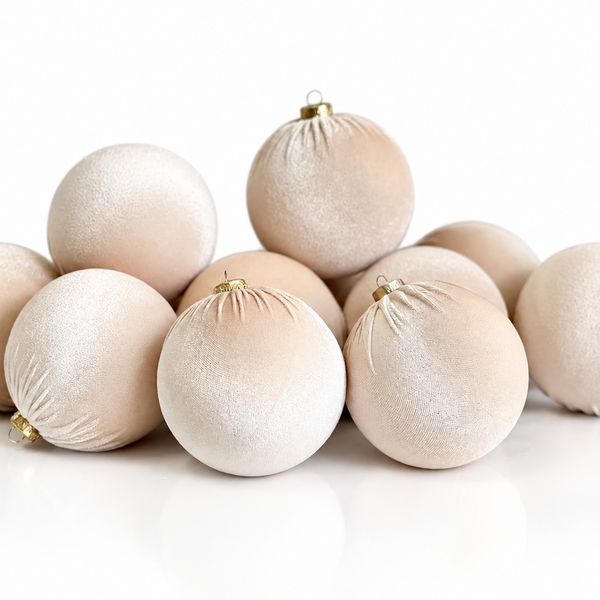 A group of matte, champagne-colored Christmas ornaments with a soft velvet-like texture and small gold hanging loops, arranged on a clean white reflective surface.