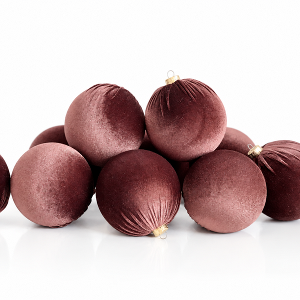 A pile of velvet Christmas ornaments in a deep burgundy color, arranged on a clean white reflective surface.