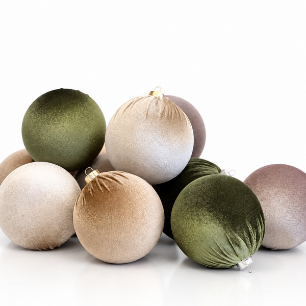 A pile of velvet Christmas baubles in muted sage green, champagne beige, and dusty mauve tones, arranged on a clean white reflective surface.