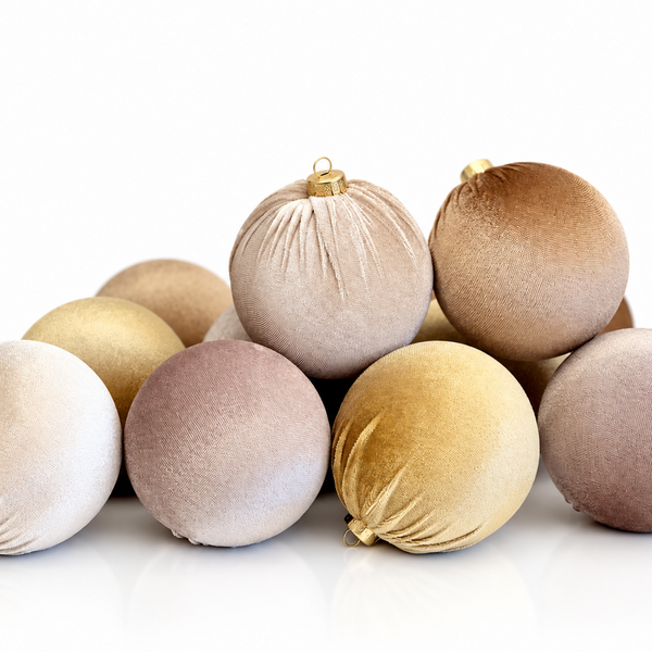 A pile of velvet Christmas ornaments in neutral shades of beige, gold, taupe, and brown, arranged on a reflective white surface against a white background.