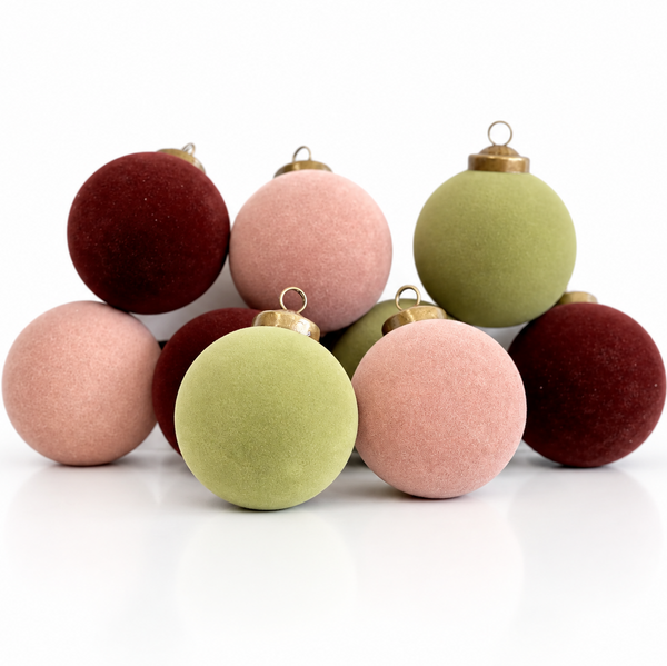 A group of nine velvet Christmas baubles in dusty pink, sage green, and deep burgundy, arranged on a reflective white surface against a white background.