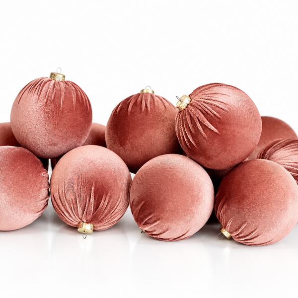 A pile of velvet Christmas ornaments in a dusty rose pink color, with small gold hanging loops, set against a clean white background.
