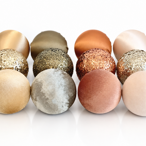 A set of 12 decorative spherical ornaments in various textures and metallic finishes, including matte, glitter, and velvet finishes in neutral and metallic colors, arranged in two rows on a white reflective surface.