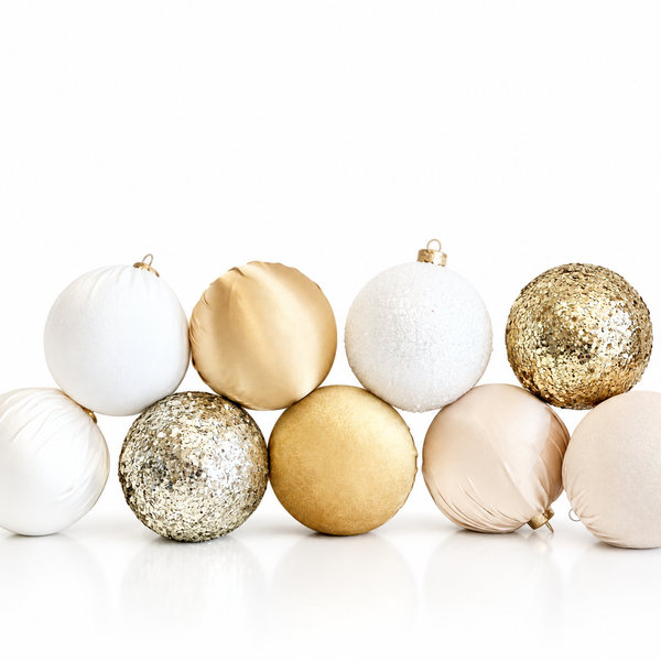 Nine decorative Christmas baubles in white, gold, and champagne beige, featuring various textures including glitter, matte, and satin, arranged on a reflective white surface against a white background.