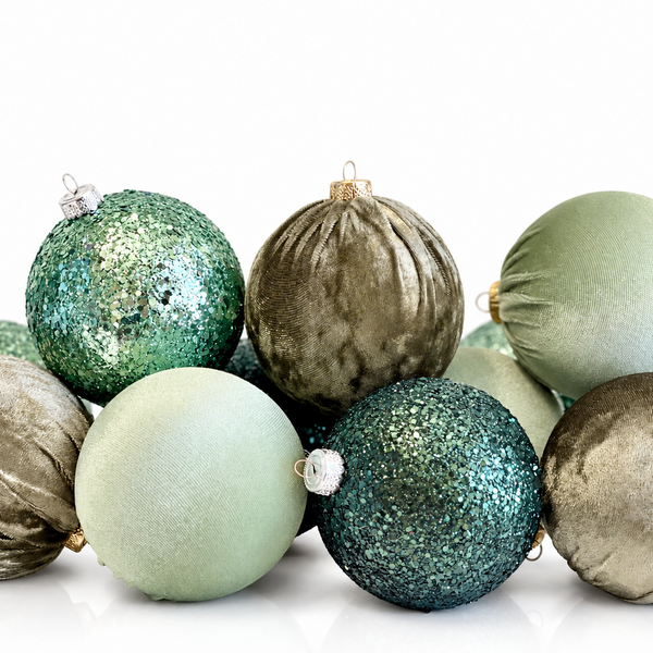 A group of decorative Christmas ornaments in sage green, teal glitter, and champagne velvet, arranged on a clean white background.