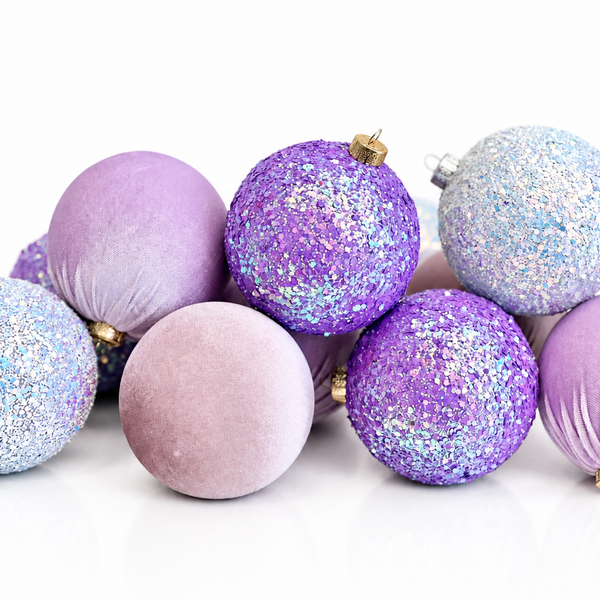 A group of decorative Christmas ornaments in shades of purple, lavender, and light blue, including both glittery and velvet finishes, arranged on a white reflective surface.