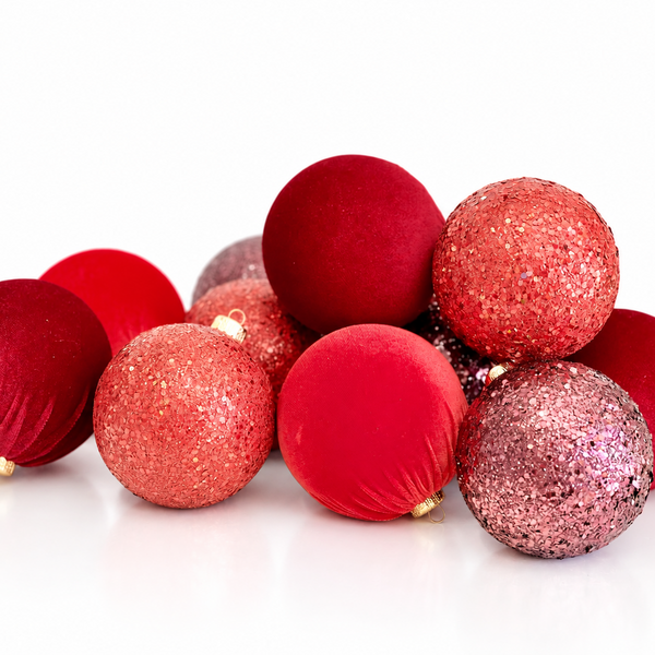A group of red and pink Christmas ornaments, including both matte velvet and glittery textured balls, arranged on a clean white background.