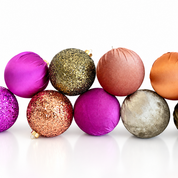 A row of decorative Christmas ornaments in assorted colors and textures, including metallic pink, glittery gold, matte copper, orange, textured silver, and deep purple, arranged on a reflective white surface against a white background.