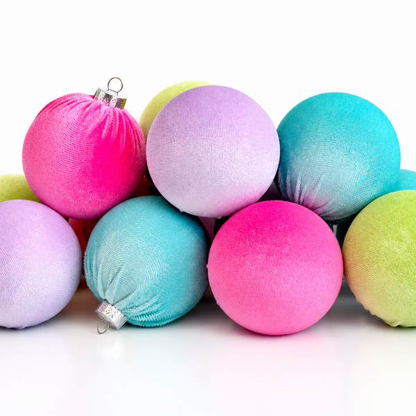A pile of colorful Christmas ornaments on a white reflective surface. The ornaments include shades of pink, purple, blue, green, and yellow, with a soft textured finish and silver hanging loops.