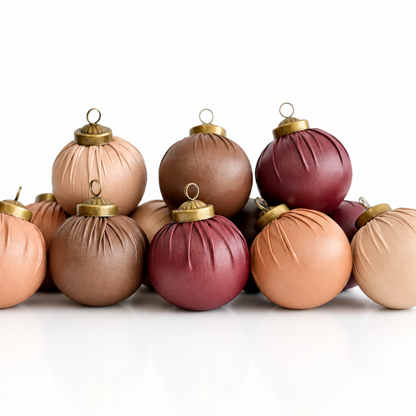 A group of fabric-covered Christmas ornaments in muted shades of terracotta, dusty rose, mauve, and peach, each with a gold metal cap and loop, arranged on a reflective white surface against a white background.